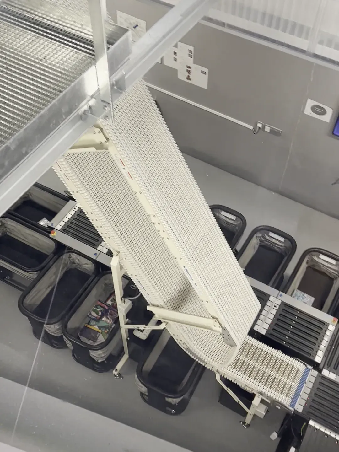 Looking down through glass at an automated book-return system in the Library of Caen, where a white modular conveyor belt curves down to drop returned books into black plastic bins